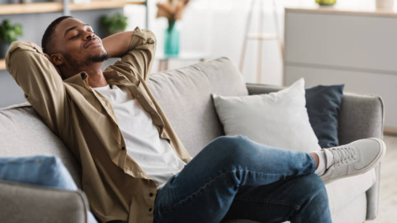 Relaxation And Comfort. Contented African American Guy Relaxing With Eyes Closed Holding Hands Behind Head Sitting On Sofa At Home. Man Enjoying Lazy Weekend In Living Room. Side View